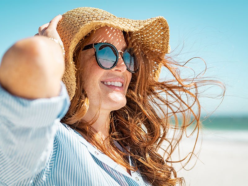 Life Extension, a smiling red-haired woman on the beach with sunglasses and a hat, wearing a blue shirt